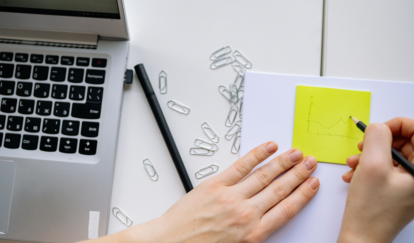 A person sketches a graph on a sticky note next to a laptop and paper clips, suggesting planning and creativity.