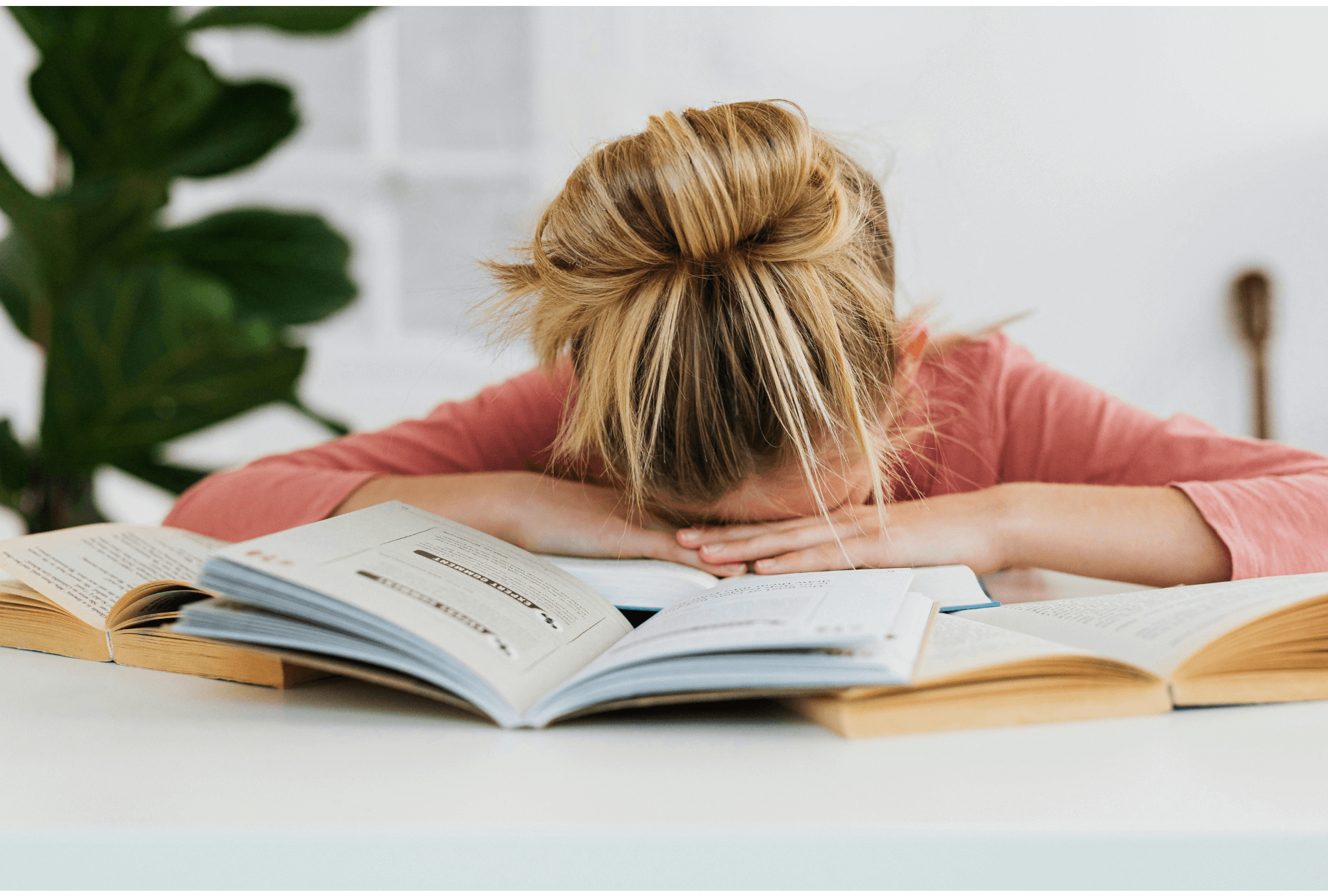 Jeune personne blonde, la tête baissée sur des livres ouverts sur une table blanche, exprimant le stress des études ou des examens.