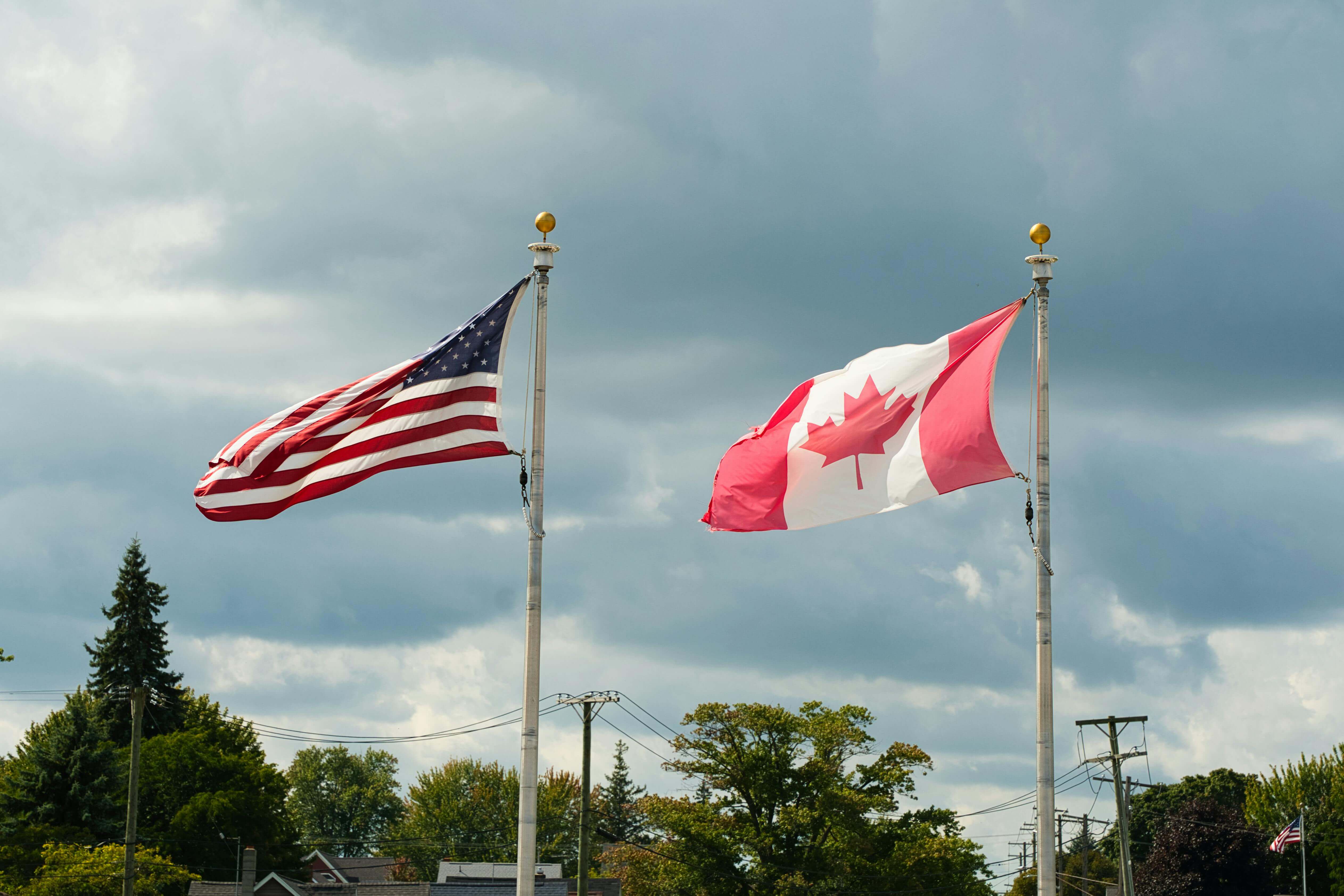 Drapeaux des États-Unis et du Canada côte à côte sous un ciel nuageux.