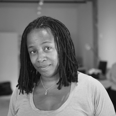 An image of Stephanie Dinkins: a black woman with dreaded hair, looking quizically at the camera, with a bluish-grey necklace and shirt.