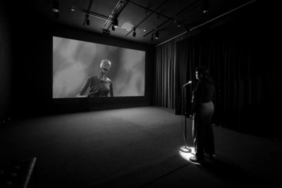 A projector in a dark room showing Rashaad Newsome's Being project: an AI represented by a 3-D rendering of a robot made of wood and metal.