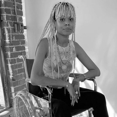 The artist, sitting in her wheelchair, looking directly at the camera. She is  a black woman with blonde cornrows and a nose ring, wearing a white shirt and dark leggings.