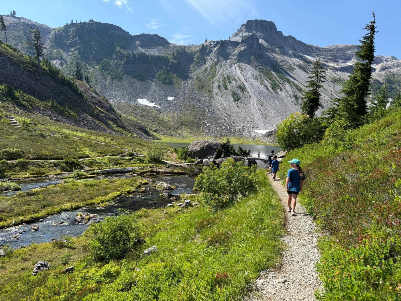 Children hiking beside a stream toward a mountain lake.
