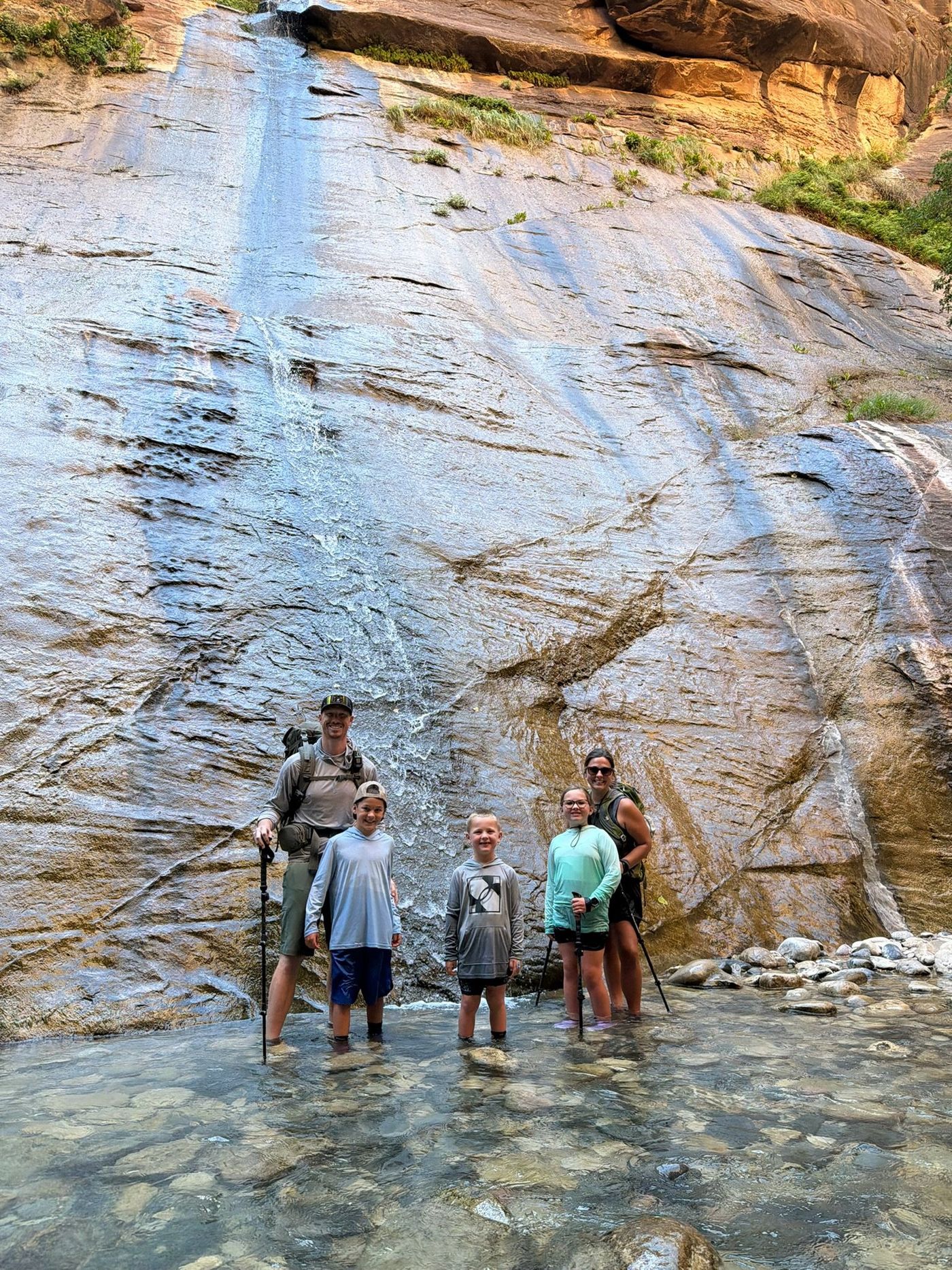 A family standing in shallow water beneath a canyon waterfall.