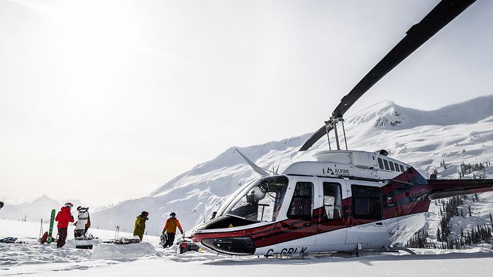 Helicopter parked on a snowy mountain plateau with several people in colourful winter gear standing nearby.