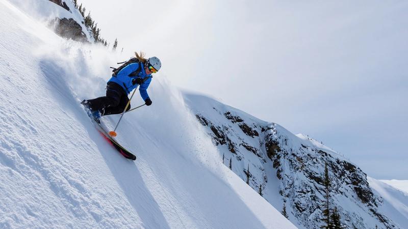 Skier in a blue jacket and black pants descending a steep, snowy mountain slope, kicking up snow with rugged, snow-covered terrain and scattered trees in the background.