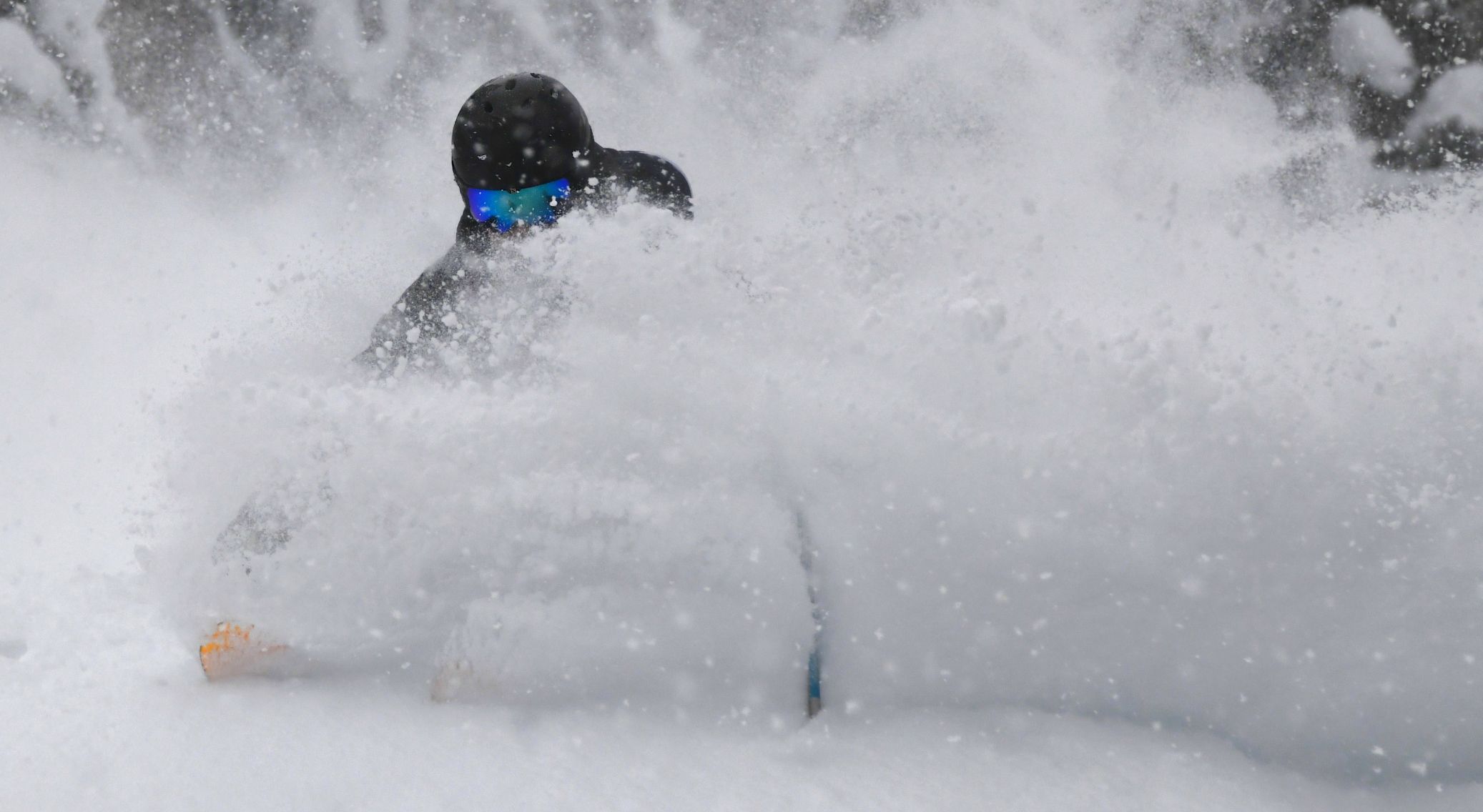Person skiing or snowboarding through deep powder snow, wearing a black helmet and blue goggles, with snow kicked up around them partially obscuring their body.