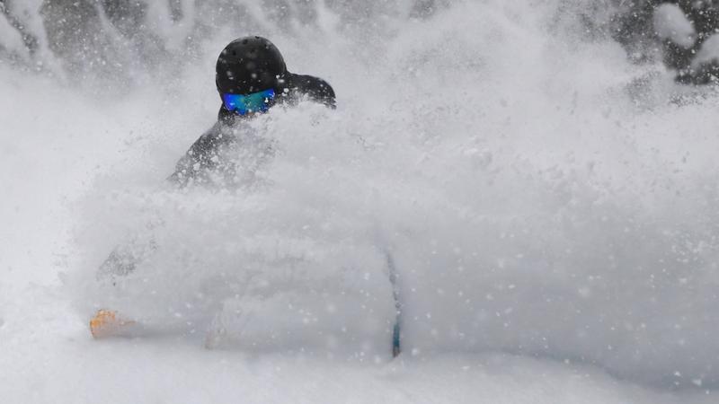 Person skiing or snowboarding through deep powder snow, wearing a black helmet and blue goggles, with snow kicked up around them partially obscuring their body.