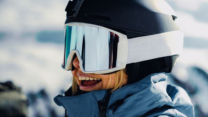 Close-up of a person wearing a black ski helmet and reflective goggles, dressed in a blue winter jacket, with a snowy mountain landscape in the background.