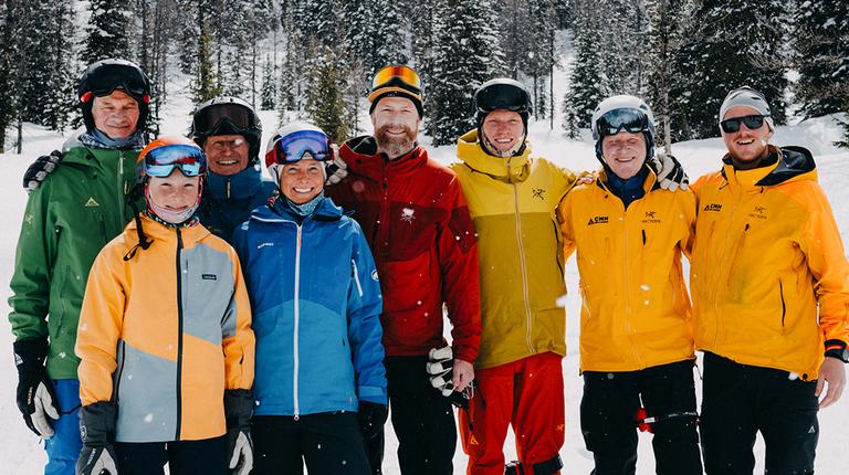 A group of eight smiling people dressed in colorful ski jackets and snow gear pose together on a snowy mountain slope surrounded by evergreen trees. They appear to be enjoying a winter ski trip, with fresh snow falling lightly around them.