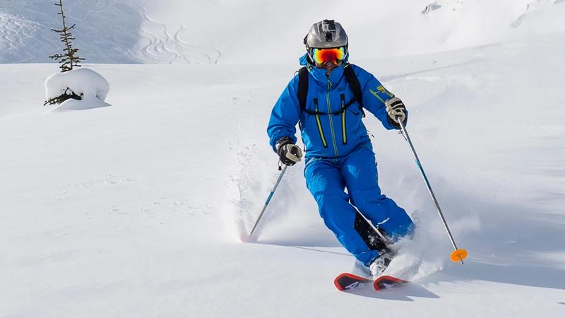 Skier in a blue ski suit, helmet, and reflective goggles skiing down a snowy slope, kicking up snow with ski poles in hand and a small snow-covered tree in the background.