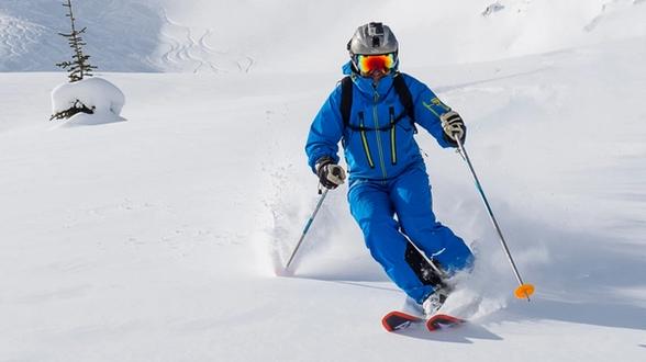 Skier in a blue ski suit, helmet, and reflective goggles skiing down a snowy slope, kicking up snow with ski poles in hand and a small snow-covered tree in the background.