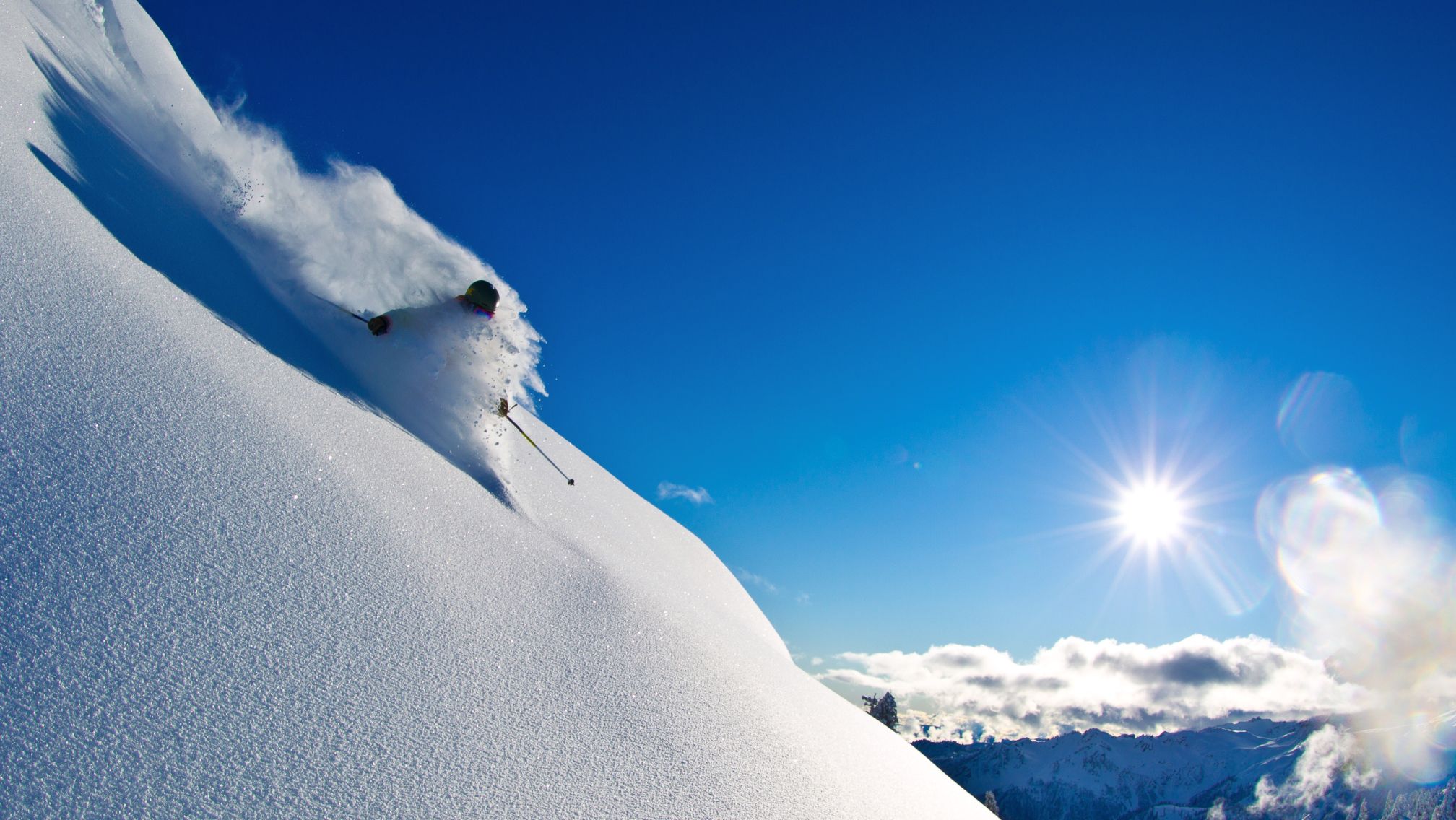 Skier descending a steep, snow-covered slope under a clear blue sky, carving through powdery snow with sunlight illuminating the landscape and distant mountain peaks in the background.