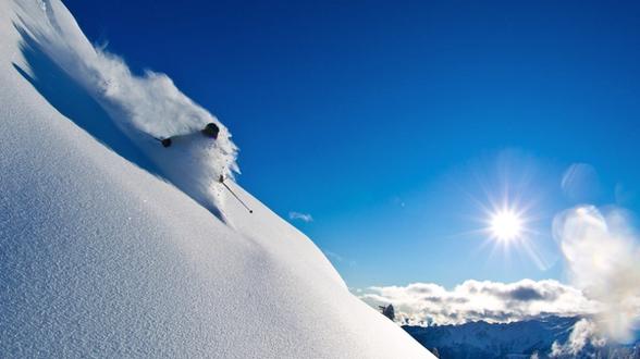 Skier descending a steep, snow-covered slope under a clear blue sky, carving through powdery snow with sunlight illuminating the landscape and distant mountain peaks in the background.