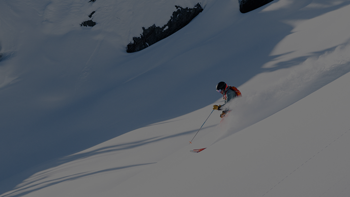 skier holding a ski pole and wearing a red bag pack sprays snow as they go down a run.