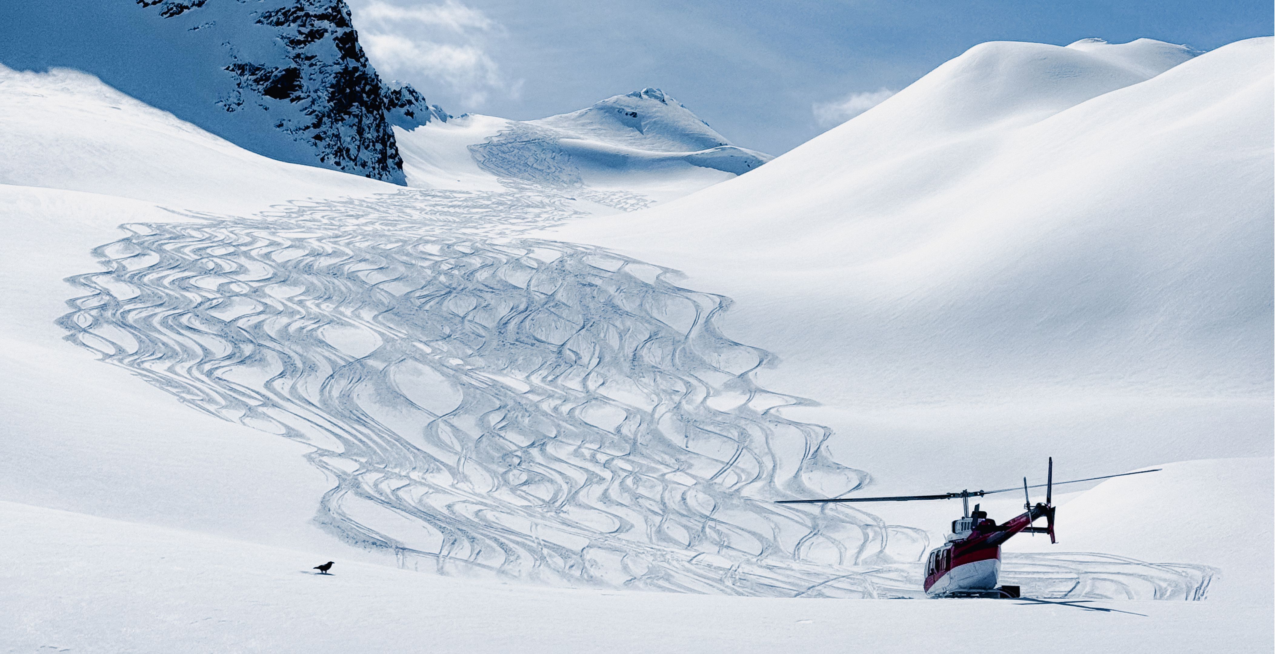 A red and white helicopter parked on a snowy mountain slope, surrounded by ski tracks under a clear blue sky, suggesting heli-skiing activity in a remote alpine landscape.