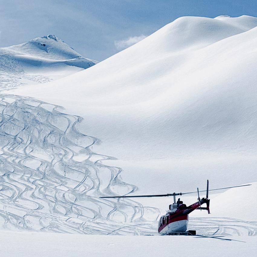 A red and white helicopter parked on a snowy mountain slope, surrounded by ski tracks under a clear blue sky, suggesting heli-skiing activity in a remote alpine landscape.