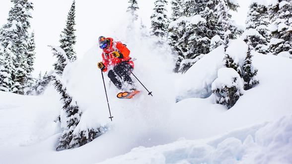 Skier in a bright orange jacket, black pants, helmet, and goggles performing a jump in mid-air with ski poles, surrounded by snow-covered trees and kicked-up snow.