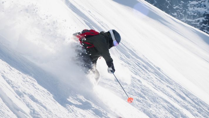 Skier carving through deep powder on a steep snowy slope, wearing a dark jacket, helmet, goggles, and a red backpack, with snow spraying up dramatically.