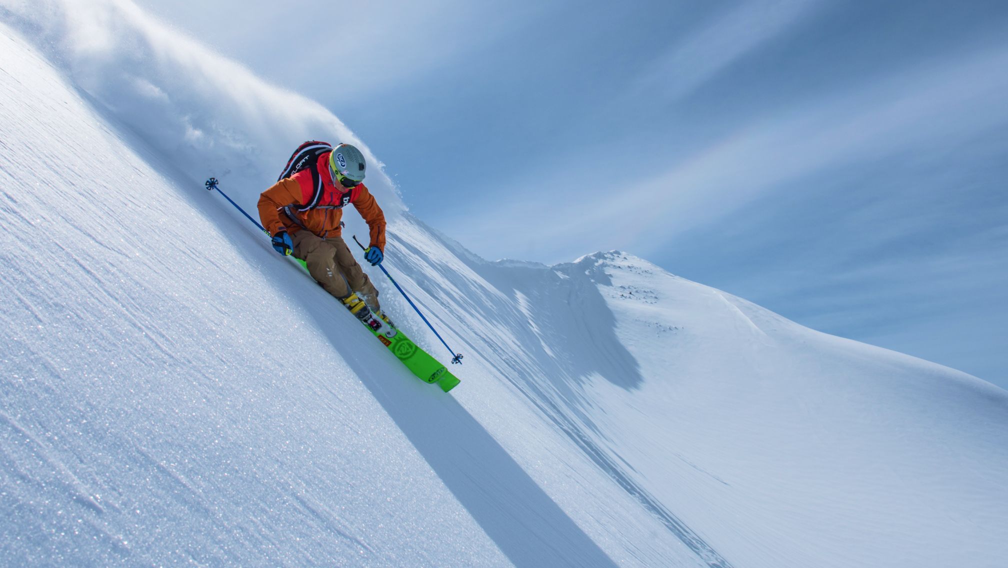 Skier in an orange jacket and brown pants descending a steep, snow-covered mountain slope under a clear blue sky, wearing a helmet, goggles, and backpack, with bright green skis and ski poles.