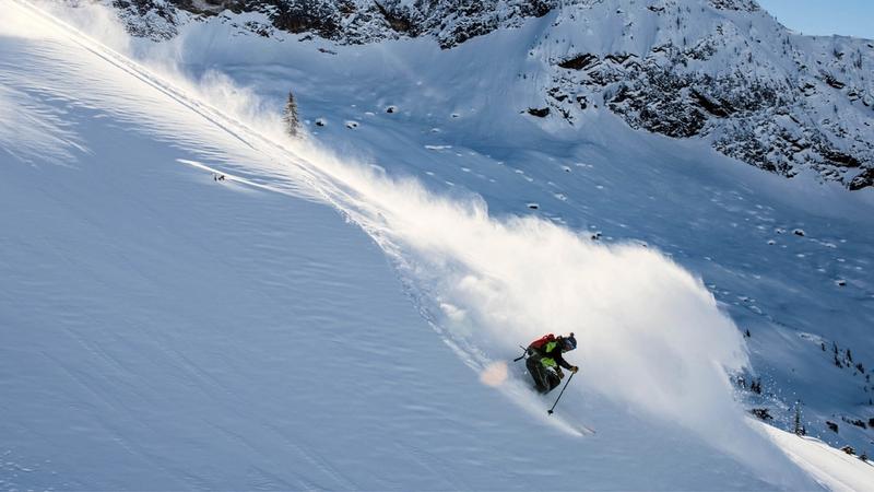 Skier making a sharp turn while descending a steep, snow-covered mountain slope, with snow spraying behind and rocky outcrops visible in the background.