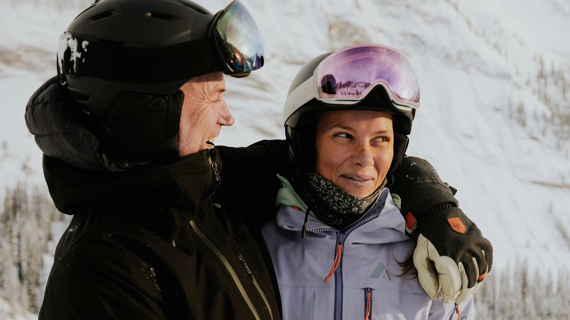 Two people dressed in winter gear and ski helmets standing close together on a snowy mountain, with one person’s arm around the other.