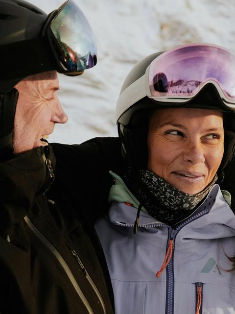 Two people dressed in winter gear and ski helmets standing close together on a snowy mountain, with one person’s arm around the other.