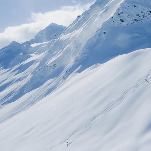 Wide snowy slope with ski tracks and skiers descending under bright sunlight.