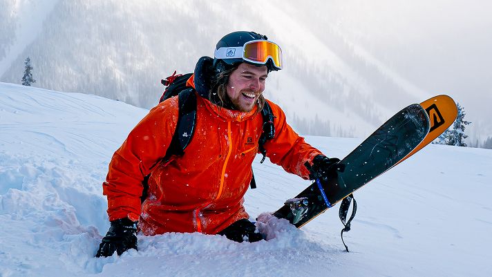 Person in a bright orange ski jacket kneeling in deep snow on a mountain slope, holding a single ski vertically.