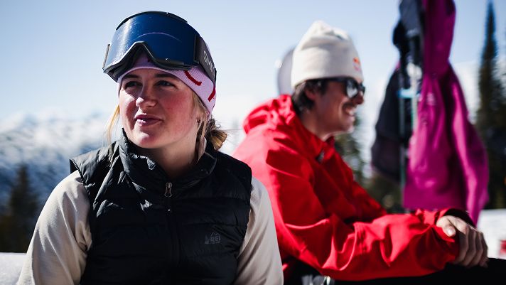 Two people in winter clothing sitting outdoors on a snowy mountain, one wearing a black helmet and vest, the other in a bright red jacket and light beanie.