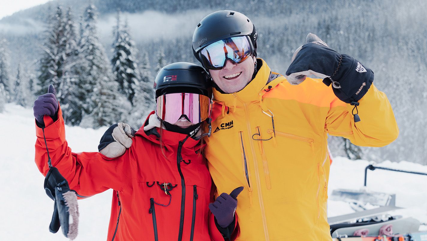 Two people in bright ski jackets and helmets posing together on a snowy mountain slope, with snow-covered trees and hills in the background.
