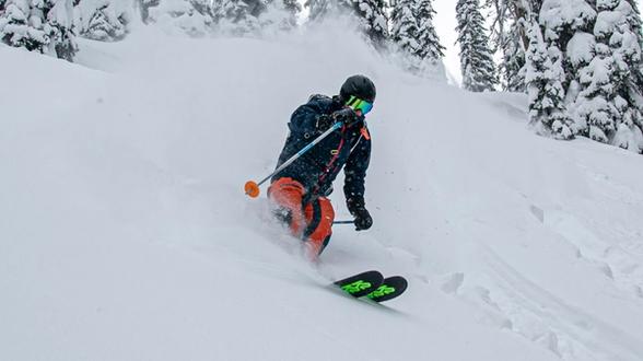 Skier in a dark helmet, reflective goggles, dark jacket, and bright orange pants skiing down a snowy slope, turning and spraying snow with snow-covered trees in the background.