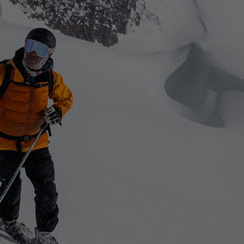 Person skiing on a snowy mountain slope, wearing a bright orange jacket, black helmet, and carrying ski poles.