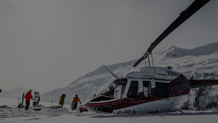 Helicopter parked on a snowy mountain plateau with several people in colourful winter gear standing nearby.