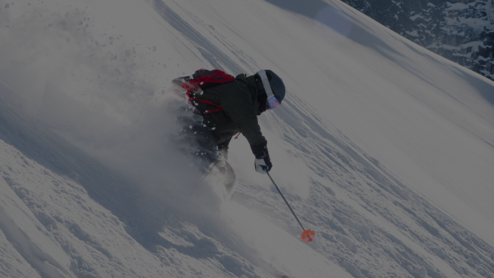 Skier carving through deep powder on a steep snowy slope, wearing a dark jacket, helmet, goggles, and a red backpack, with snow spraying up dramatically.