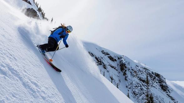 Skier in a blue jacket and black pants descending a steep, snowy mountain slope, kicking up snow with rugged, snow-covered terrain and scattered trees in the background.