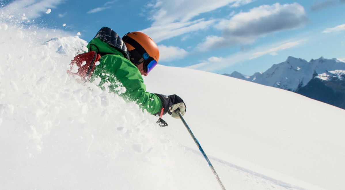 skier in a green jacket carves a powerful turn exploding through untouched powder with sharp mountain peaks visible in the distance on a cmh heli-skiing adventure