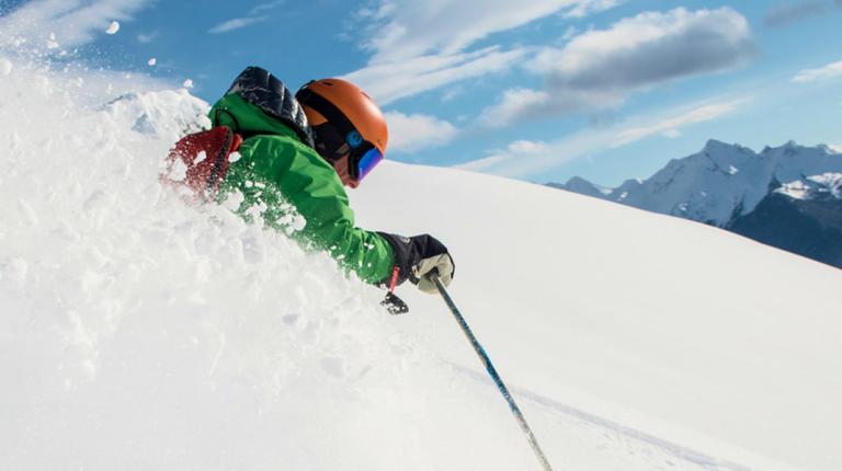 skier in a green jacket carves a powerful turn exploding through untouched powder with sharp mountain peaks visible in the distance on a cmh heli-skiing adventure