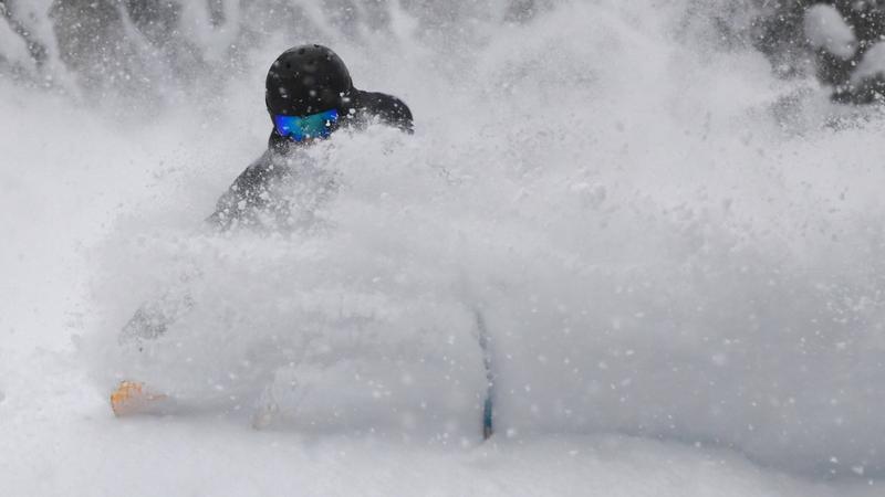 Person skiing or snowboarding through deep powder snow, wearing a black helmet and blue goggles, with snow kicked up around them partially obscuring their body.