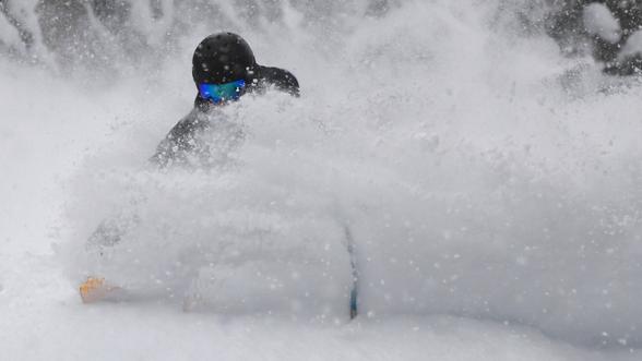 Person skiing or snowboarding through deep powder snow, wearing a black helmet and blue goggles, with snow kicked up around them partially obscuring their body.