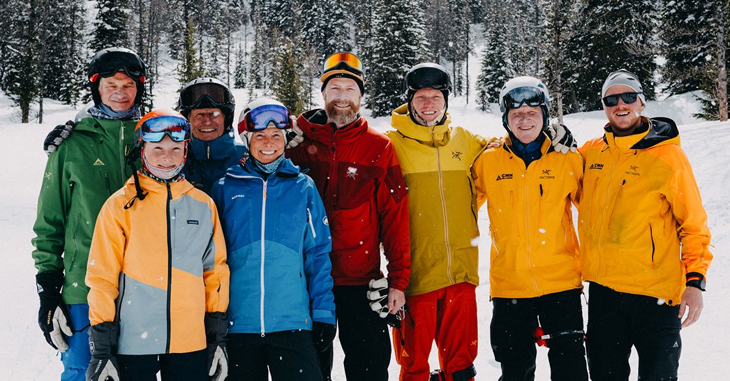 A group of eight smiling people dressed in colorful ski jackets and snow gear pose together on a snowy mountain slope surrounded by evergreen trees. They appear to be enjoying a winter ski trip, with fresh snow falling lightly around them.