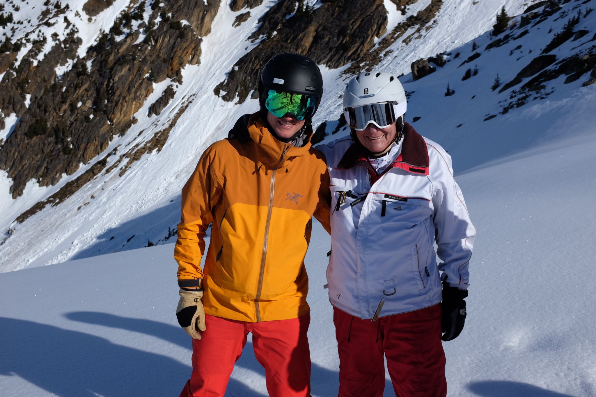 two happy skiers pose together against a dramatic snowy rocky background celebrating an incredible day of remote mountain adventure with cmh heli-skiing