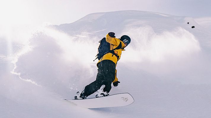 Person snowboarding down a slope in deep powder, wearing a bright yellow jacket and black helmet, with snow spraying up dramatically behind them against a backdrop of snowy mountains.