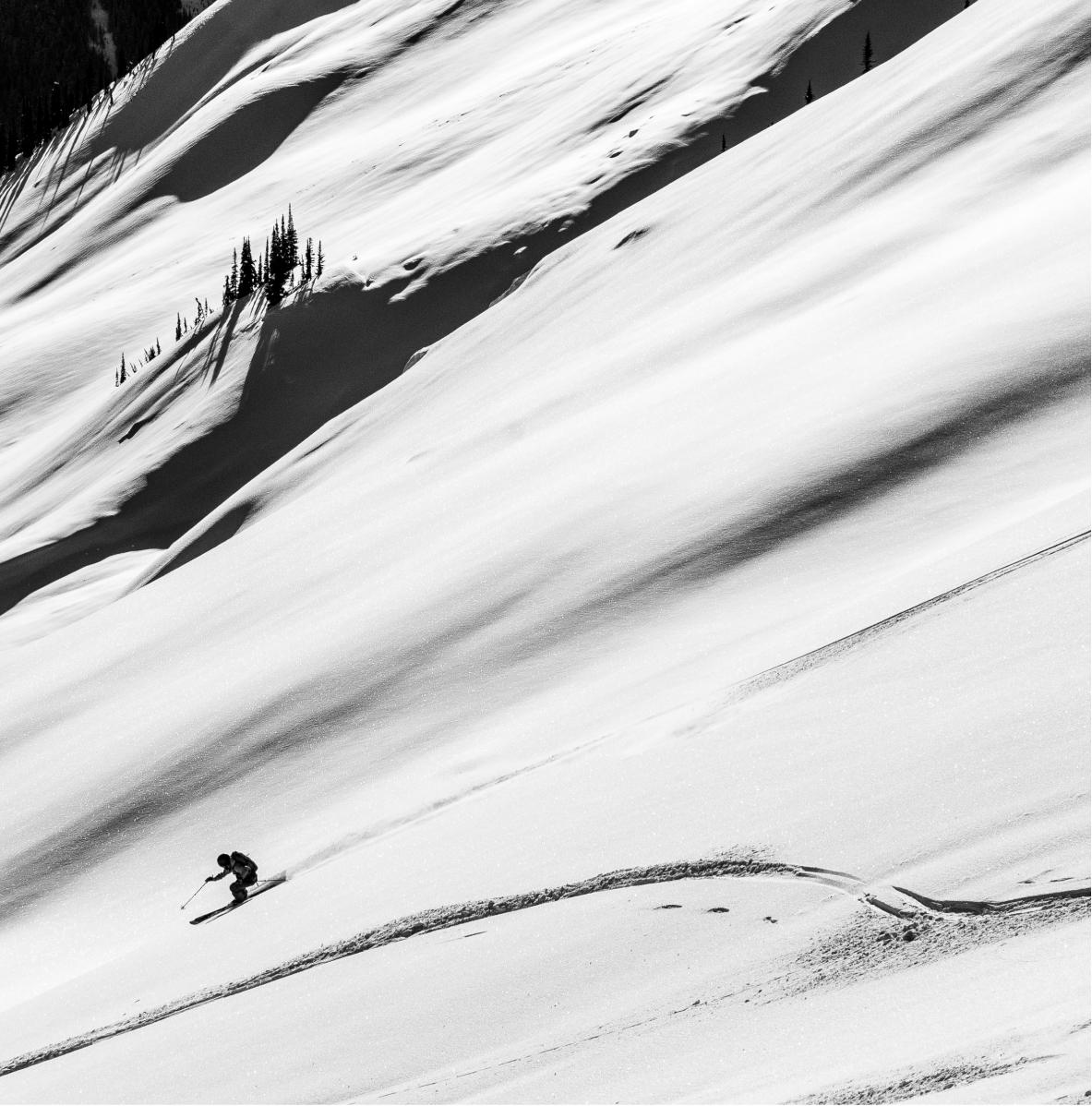 a dramatic black and white image of a lone skier making large sweeping turns down a massive steep untracked powder field highlighting the isolation and adventure of cmh heli-skiing