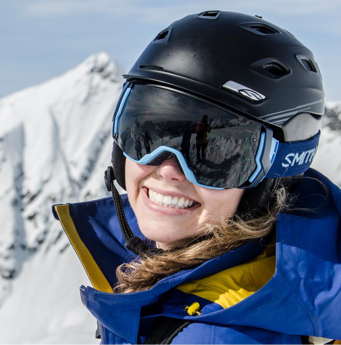 a happy skier skier smiles widely in her helmet and goggles standing on a snowy peak with sharp mountains in the background during her cmh heli-skiing trip