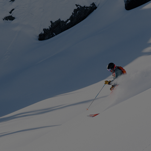 skier holding a ski pole and wearing a red bag pack sprays snow as they go down a run.
