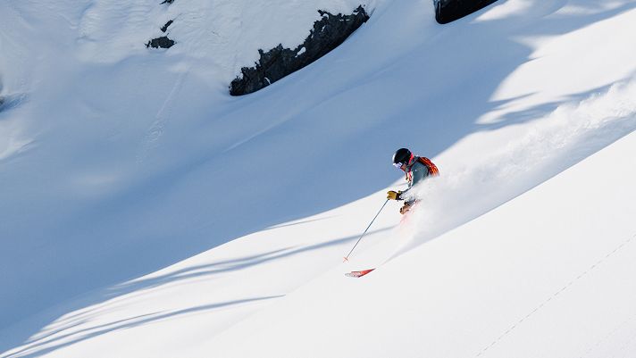 skier holding a ski pole and wearing a red bag pack sprays snow as they go down a run.