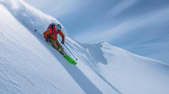 Skier in an orange jacket and brown pants descending a steep, snow-covered mountain slope under a clear blue sky, wearing a helmet, goggles, and backpack, with bright green skis and ski poles.
