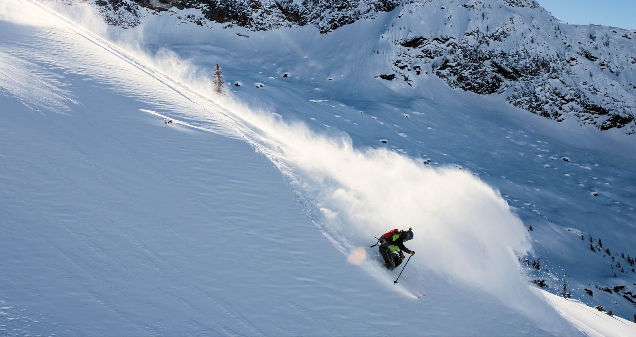 Skier making a sharp turn while descending a steep, snow-covered mountain slope, with snow spraying behind and rocky outcrops visible in the background.