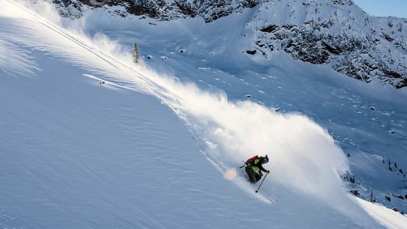 Skier making a sharp turn while descending a steep, snow-covered mountain slope, with snow spraying behind and rocky outcrops visible in the background.